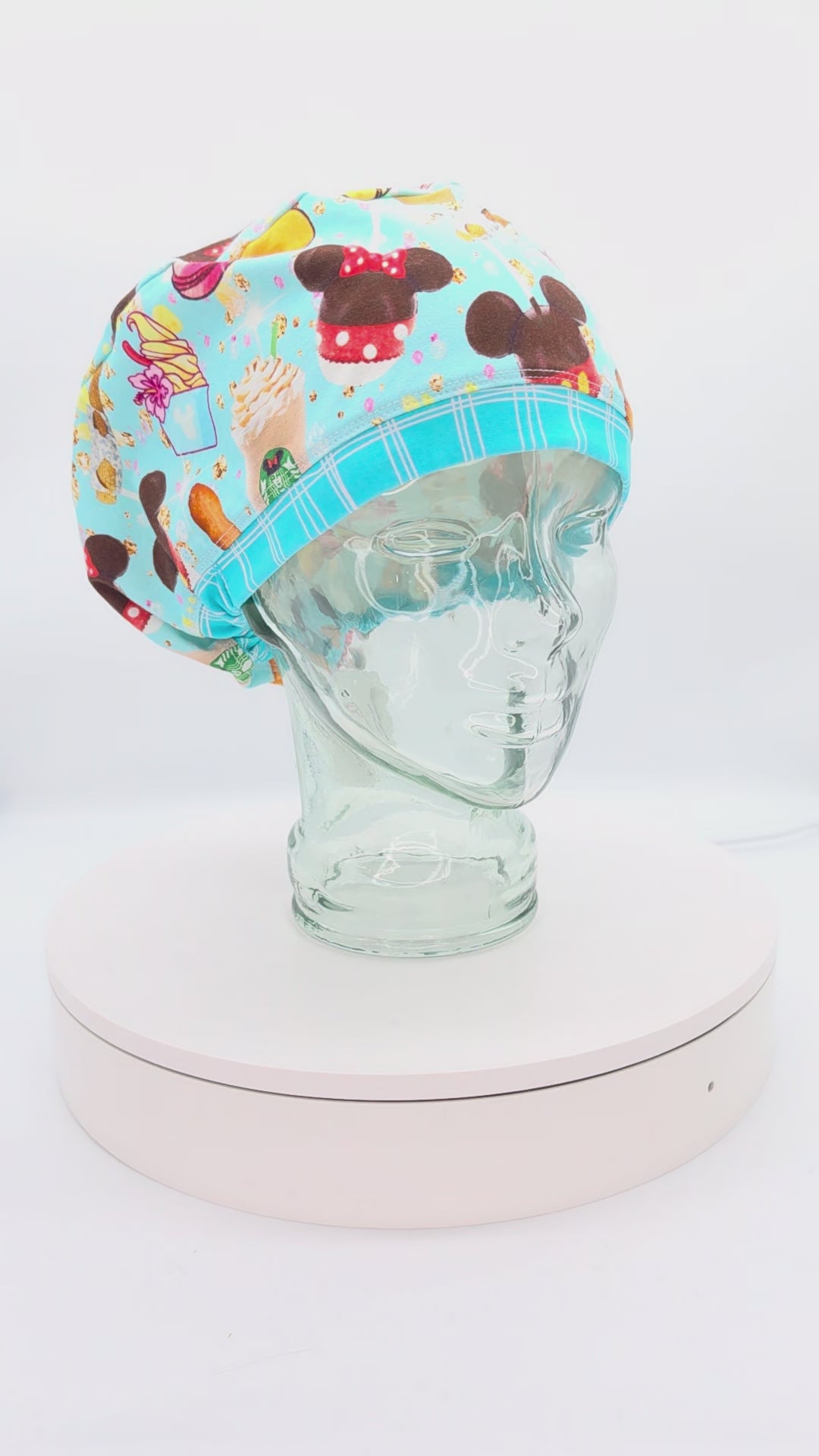 A 14-second rotating product video showing a 360-degree view of the Magical Snacks Full Coverage Scrub Cap on a clear mannequin head. The medical hat features a vibrant light blue fabric patterned with theme park treats like popcorn buckets, churros, soft pretzels, hot dogs, and mouse-shaped ice cream bars, finished with a contrasting blue-and-white striped brim.