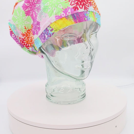 A 14-second rotating product video providing a 360-degree view of the Colorful Snowflakes Full Coverage Scrub Cap on a clear mannequin head. The medical hat features a vibrant pink fabric densely patterned with intricate, multi-colored geometric snowflakes in shades of neon green, bright orange, teal, and purple, finished with a contrasting bright yellow brim.