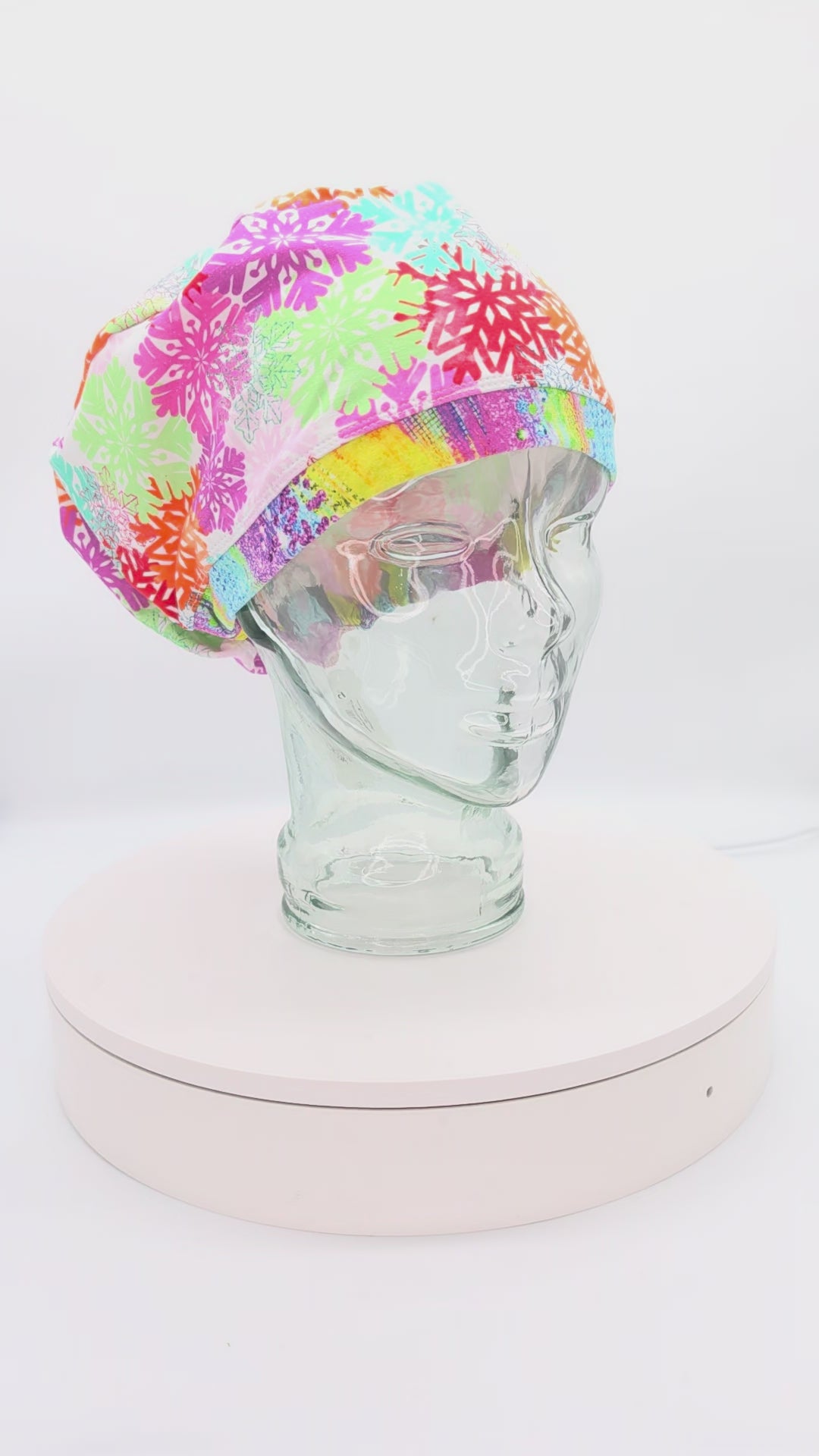 A 14-second rotating product video providing a 360-degree view of the Colorful Snowflakes Full Coverage Scrub Cap on a clear mannequin head. The medical hat features a vibrant pink fabric densely patterned with intricate, multi-colored geometric snowflakes in shades of neon green, bright orange, teal, and purple, finished with a contrasting bright yellow brim.