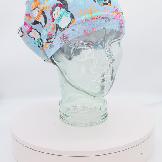 A 360-degree rotation video of a clear glass mannequin head on a white spinning display turntable, showcasing a light blue Christmas Penguins scrub cap to demonstrate its full-coverage design and adjustable cord lock toggle fit from all angles.