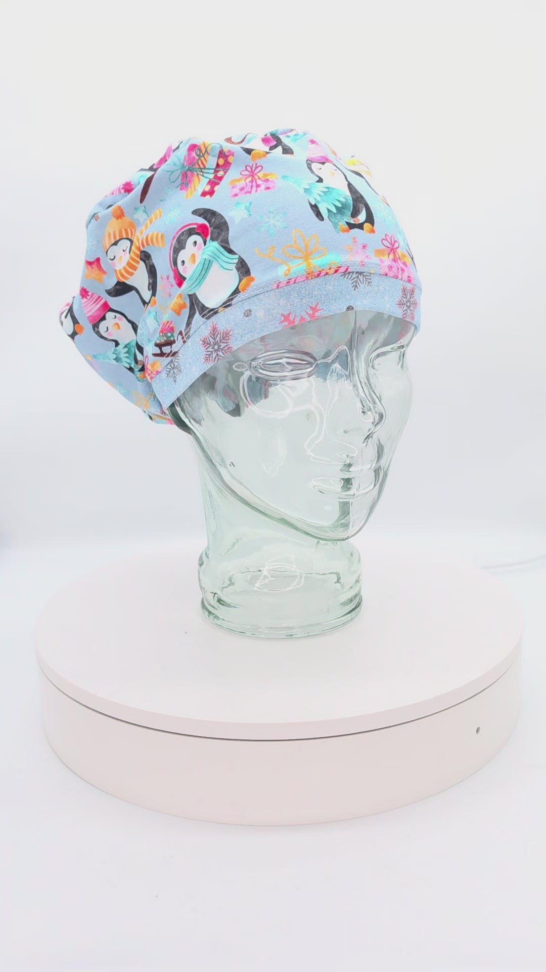 A 360-degree rotation video of a clear glass mannequin head on a white spinning display turntable, showcasing a light blue Christmas Penguins scrub cap to demonstrate its full-coverage design and adjustable cord lock toggle fit from all angles.