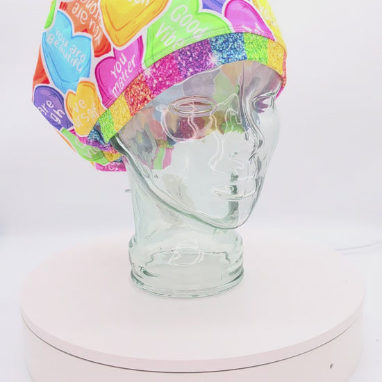 A 360-degree rotation video of a clear glass mannequin head on a white spinning display turntable, showcasing a colorful affirmation hearts scrub cap to demonstrate its full-coverage design and adjustable cord lock toggle fit from all angles.