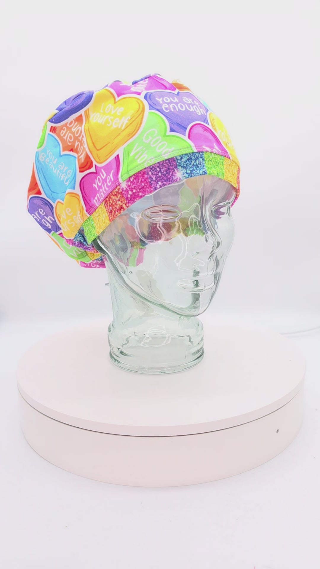 A 360-degree rotation video of a clear glass mannequin head on a white spinning display turntable, showcasing a colorful affirmation hearts scrub cap to demonstrate its full-coverage design and adjustable cord lock toggle fit from all angles.