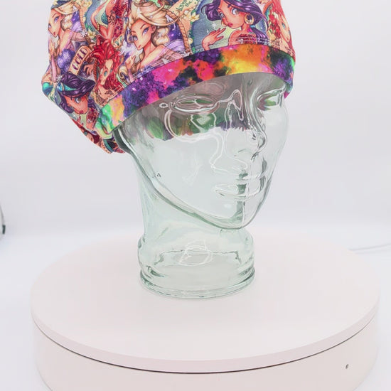 A 360-degree rotating view of a Tattoo Princess full-coverage Euro-style scrub cap on a clear mannequin head. The premium cotton knit fabric features an edgy royal design with colorful illustrations on a clean white background.
