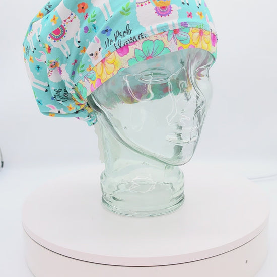 A 360-degree rotating video showcase of the "No Prob Llama" full-coverage scrub cap on a clear mannequin head, displaying its vibrant blue and white llama pattern with floral accents, stretchy cotton-elastane fabric, and adjustable elastic back with a heavy-duty cord lock toggle.