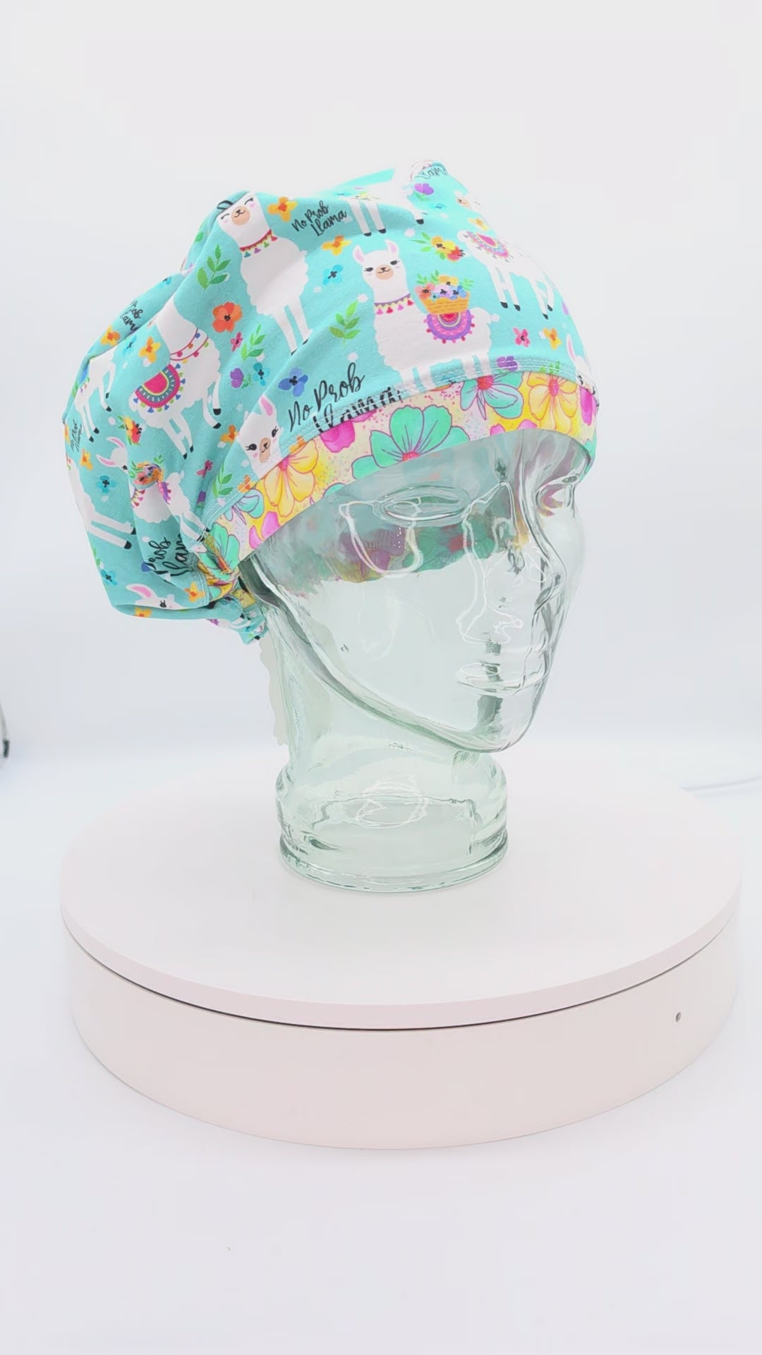 A 360-degree rotating video showcase of the "No Prob Llama" full-coverage scrub cap on a clear mannequin head, displaying its vibrant blue and white llama pattern with floral accents, stretchy cotton-elastane fabric, and adjustable elastic back with a heavy-duty cord lock toggle.