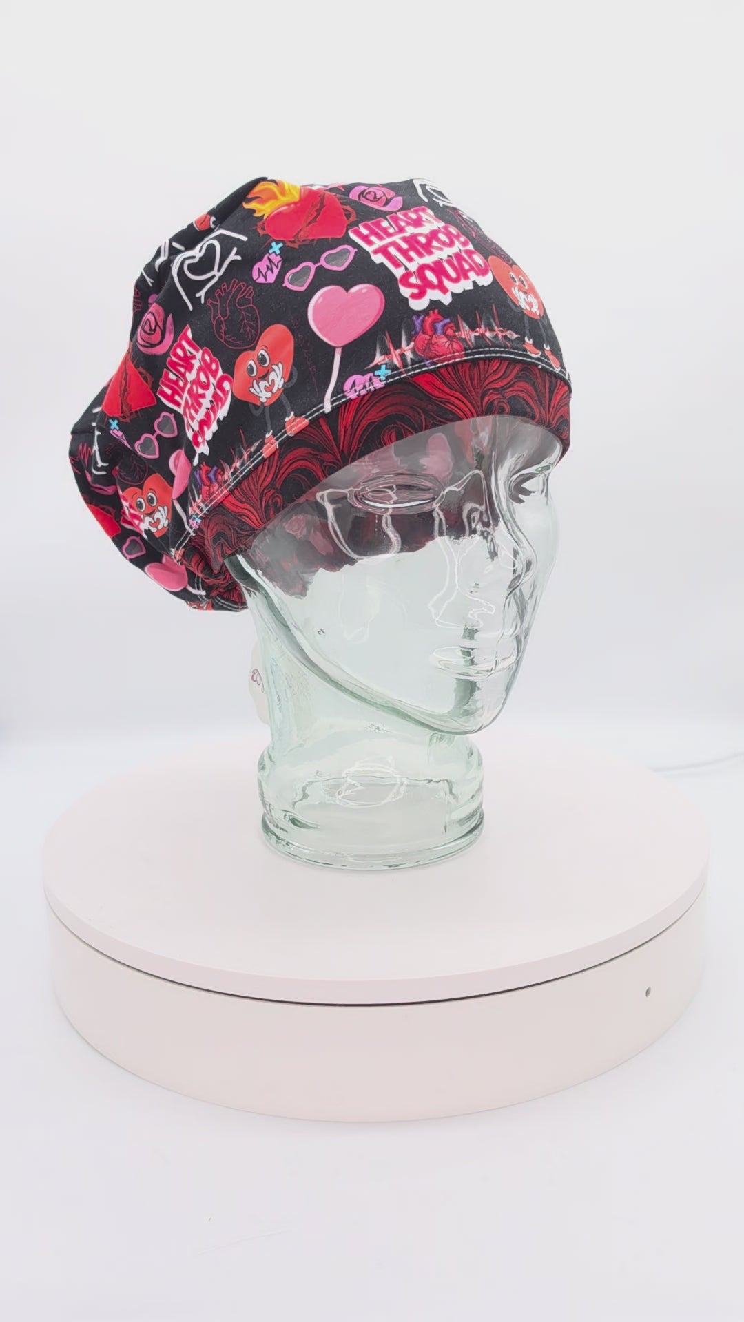 360-degree video showcase of the Heart Throb Squad scrub cap on a clear mannequin head, displaying the vibrant red, black, and pink cardiology-themed anatomical heart pattern and full-coverage fit.