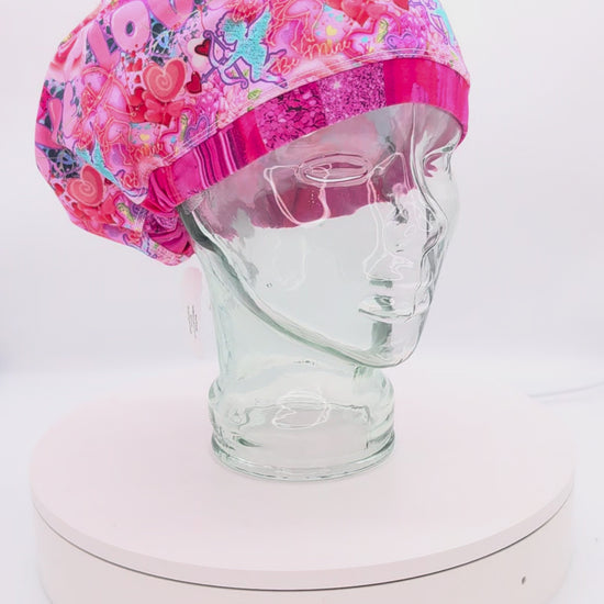 Video showcasing a 360-degree rotating view of the Cupids Love Scrub Cap on a mannequin head, highlighting the vibrant pink and red heart pattern and adjustable toggle fit.