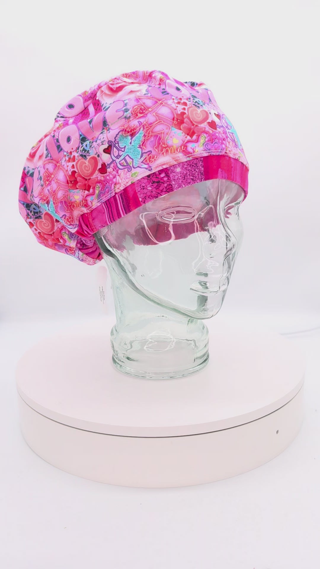 Video showcasing a 360-degree rotating view of the Cupids Love Scrub Cap on a mannequin head, highlighting the vibrant pink and red heart pattern and adjustable toggle fit.