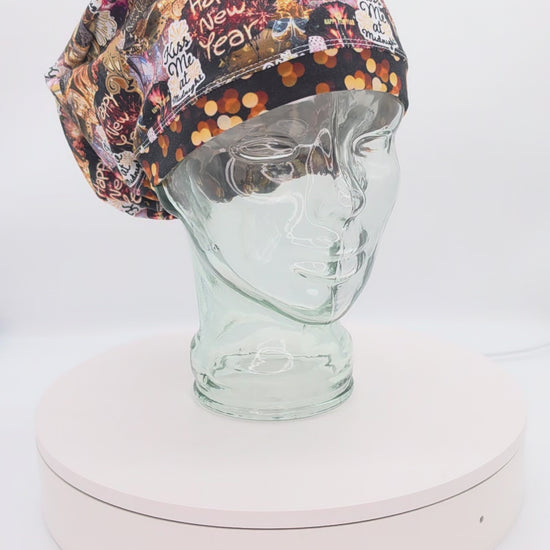 A 360-degree rotation video of a clear glass mannequin head on a white spinning display turntable, showcasing a black New Year Party scrub cap with a vibrant, multi-colored pattern of holiday clocks and celebration motifs to demonstrate its full-coverage design and adjustable cord lock toggle fit from all angles.
