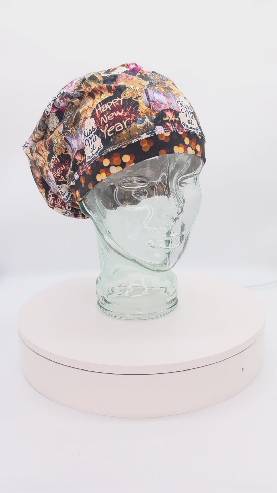 A 360-degree rotation video of a clear glass mannequin head on a white spinning display turntable, showcasing a black New Year Party scrub cap with a vibrant, multi-colored pattern of holiday clocks and celebration motifs to demonstrate its full-coverage design and adjustable cord lock toggle fit from all angles.