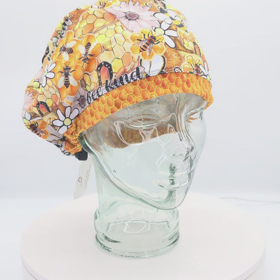 A 360-degree rotating view of a full coverage medical scrub cap on a clear mannequin head, featuring a vibrant "Bee Kind" print with honeycomb patterns, sunflowers, and honeybees.