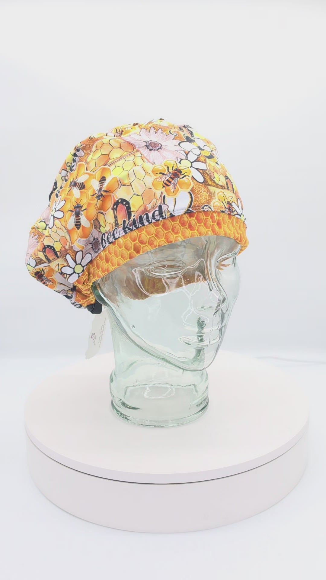 A 360-degree rotating view of a full coverage medical scrub cap on a clear mannequin head, featuring a vibrant "Bee Kind" print with honeycomb patterns, sunflowers, and honeybees.