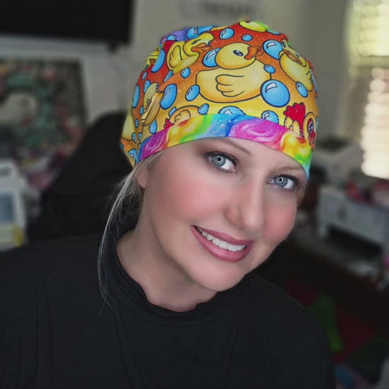 A smiling female healthcare worker wearing a vibrant, full coverage medical scrub cap featuring a custom pattern of yellow rubber ducks and blue bubbles.