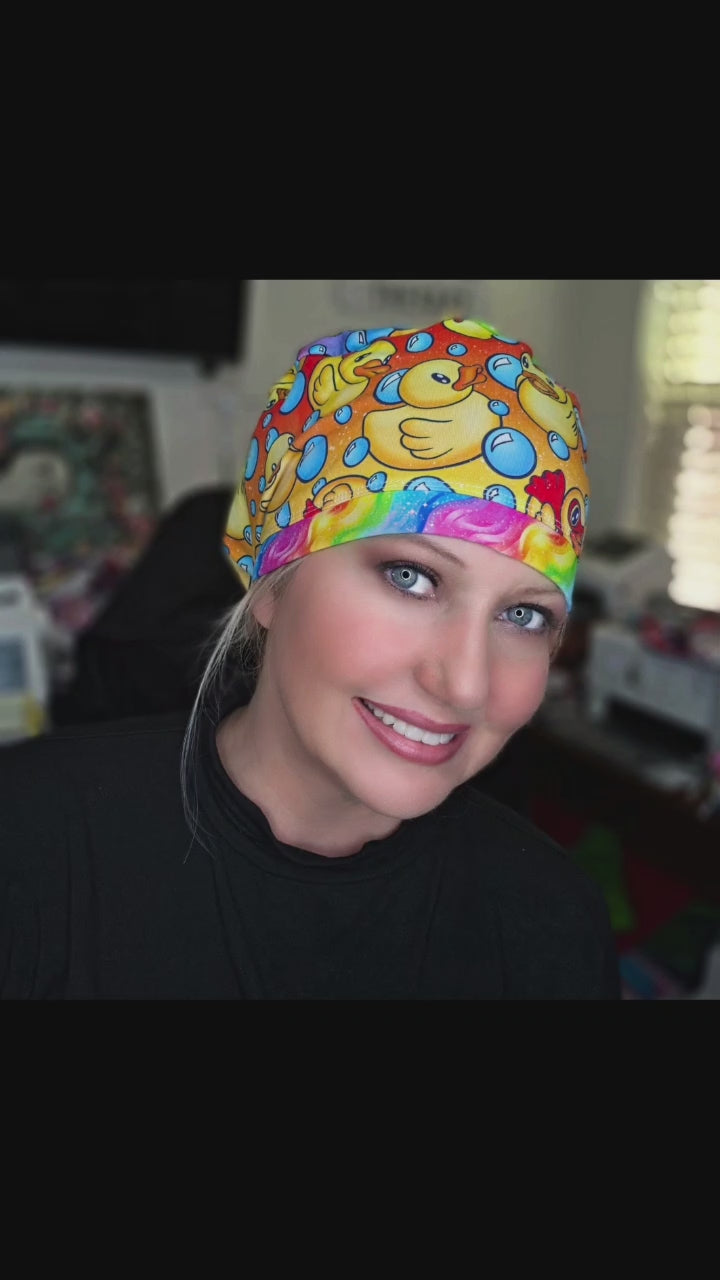 A smiling female healthcare worker wearing a vibrant, full coverage medical scrub cap featuring a custom pattern of yellow rubber ducks and blue bubbles.