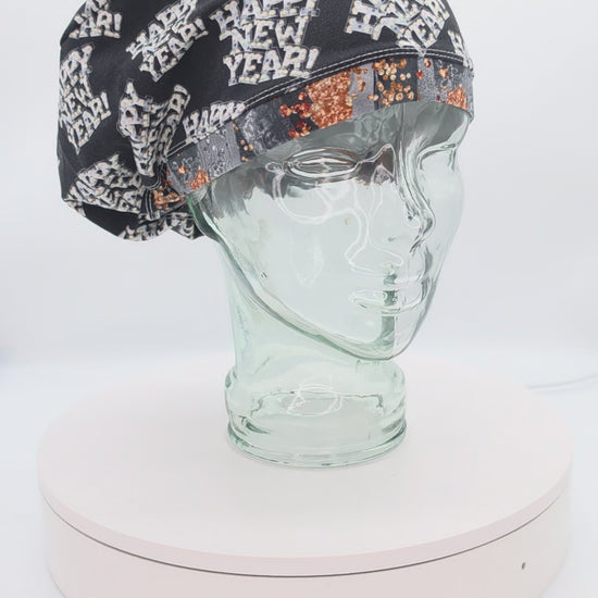 A 360-degree rotation video of a clear glass mannequin head on a white spinning display turntable, showcasing a black Happy New Year scrub cap with a white and gold festive print to demonstrate its full-coverage design and adjustable cord lock toggle fit from all angles.
