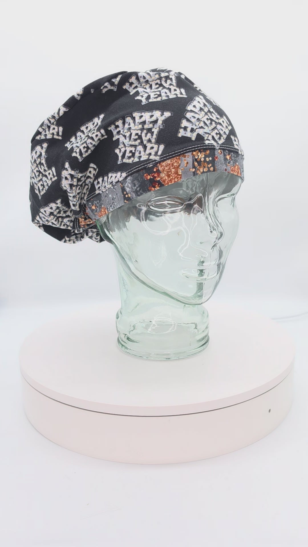A 360-degree rotation video of a clear glass mannequin head on a white spinning display turntable, showcasing a black Happy New Year scrub cap with a white and gold festive print to demonstrate its full-coverage design and adjustable cord lock toggle fit from all angles.