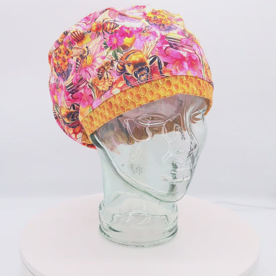 A 360-degree rotating view of a full coverage medical scrub cap on a clear mannequin head, featuring a vibrant orange and yellow bumblebee and honeycomb pattern.