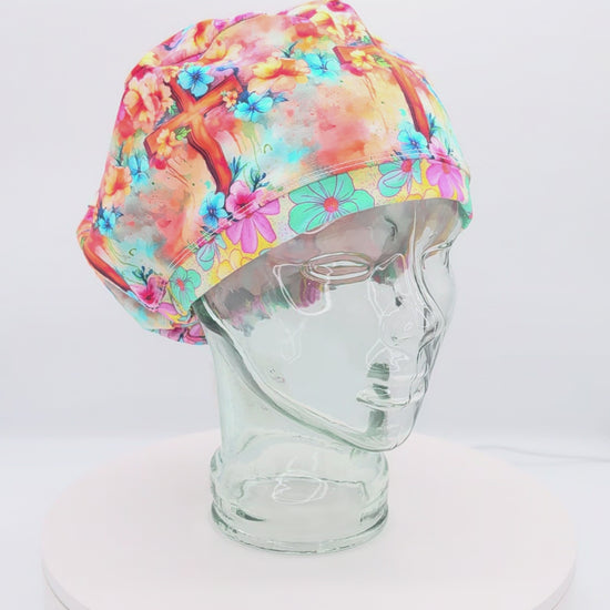 360-degree video preview of the Watercolor Cross Scrub Cap on a clear mannequin head, showcasing the colorful floral and watercolor cross pattern, euro-style fit, and adjustable back toggle.