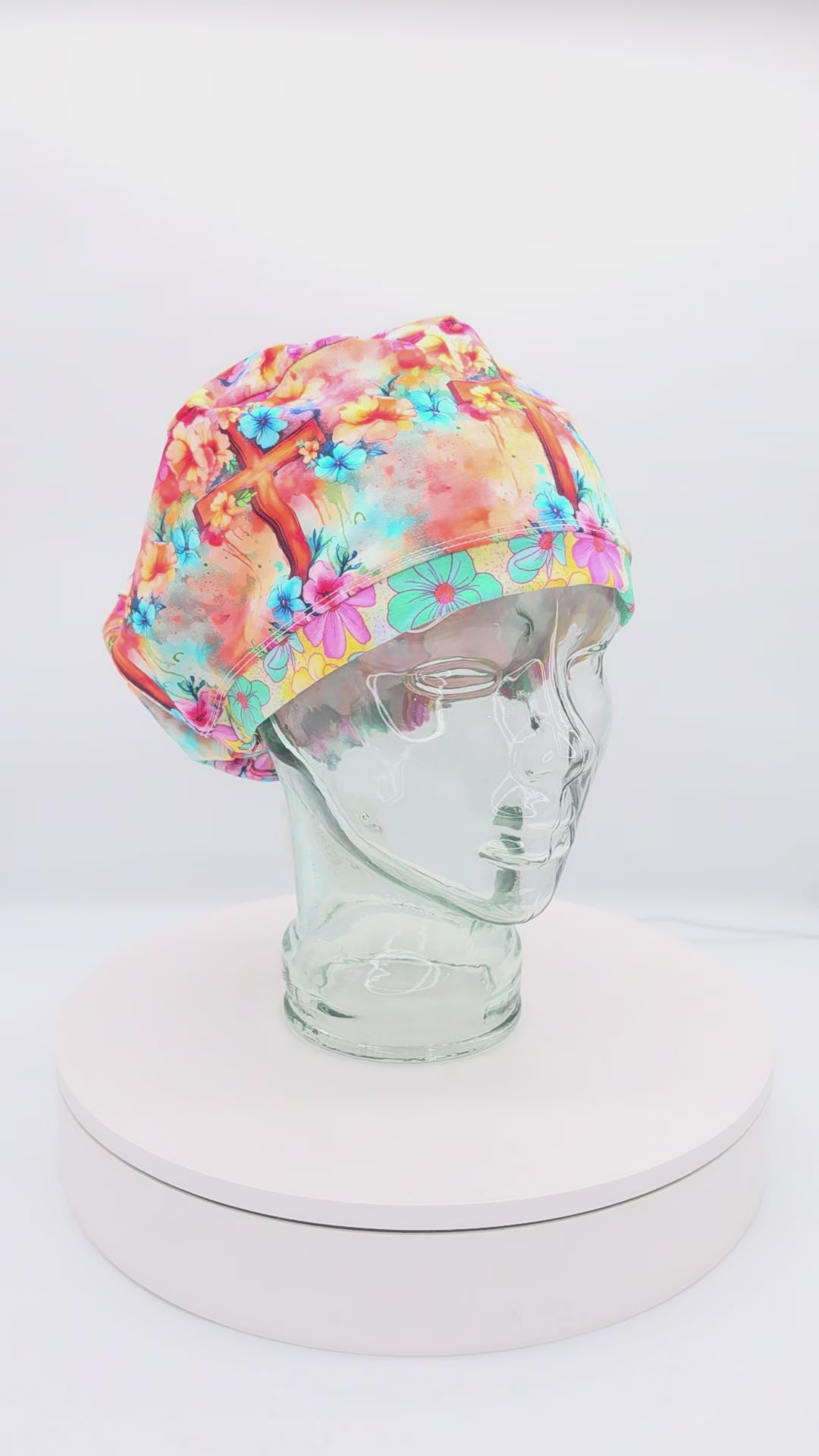 360-degree video preview of the Watercolor Cross Scrub Cap on a clear mannequin head, showcasing the colorful floral and watercolor cross pattern, euro-style fit, and adjustable back toggle.
