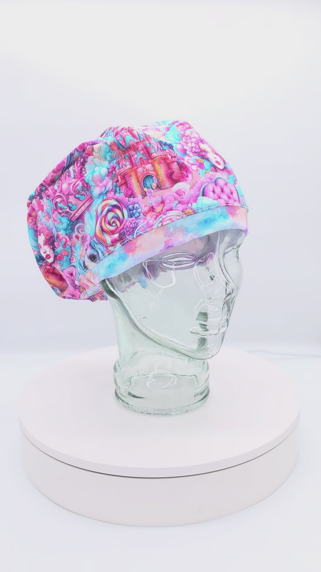 A 360-degree rotating view of a Full Coverage Scrub Cap on a clear mannequin head, displaying a vibrant, colorful pastel candy fairytale pattern with cotton candy clouds and whimsical sweets.