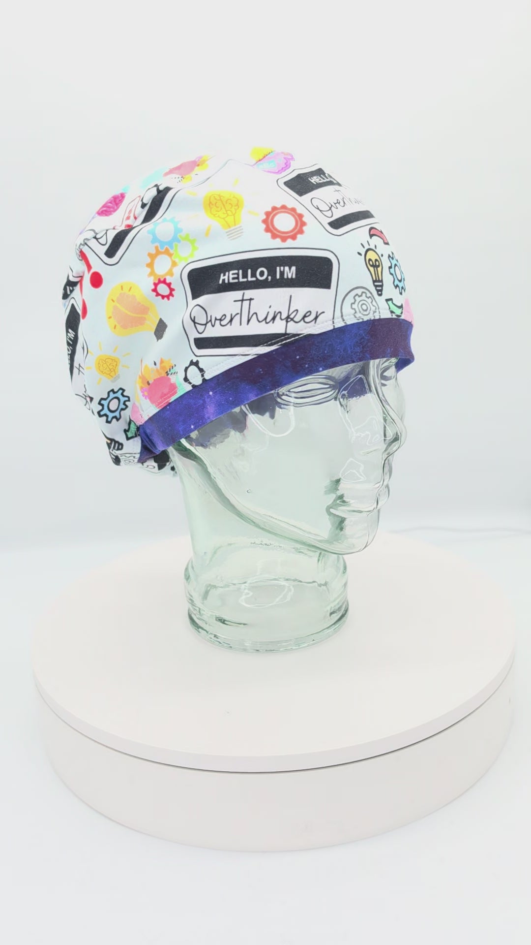 A 360-degree rotating product video showcasing the 'Overthinker' full-coverage scrub cap on a clear display stand, highlighting the colorful comic-book style graphics, 'Hello, I'm Overthinker' name tag badge illustration, and the secure, snug fit around the brim.