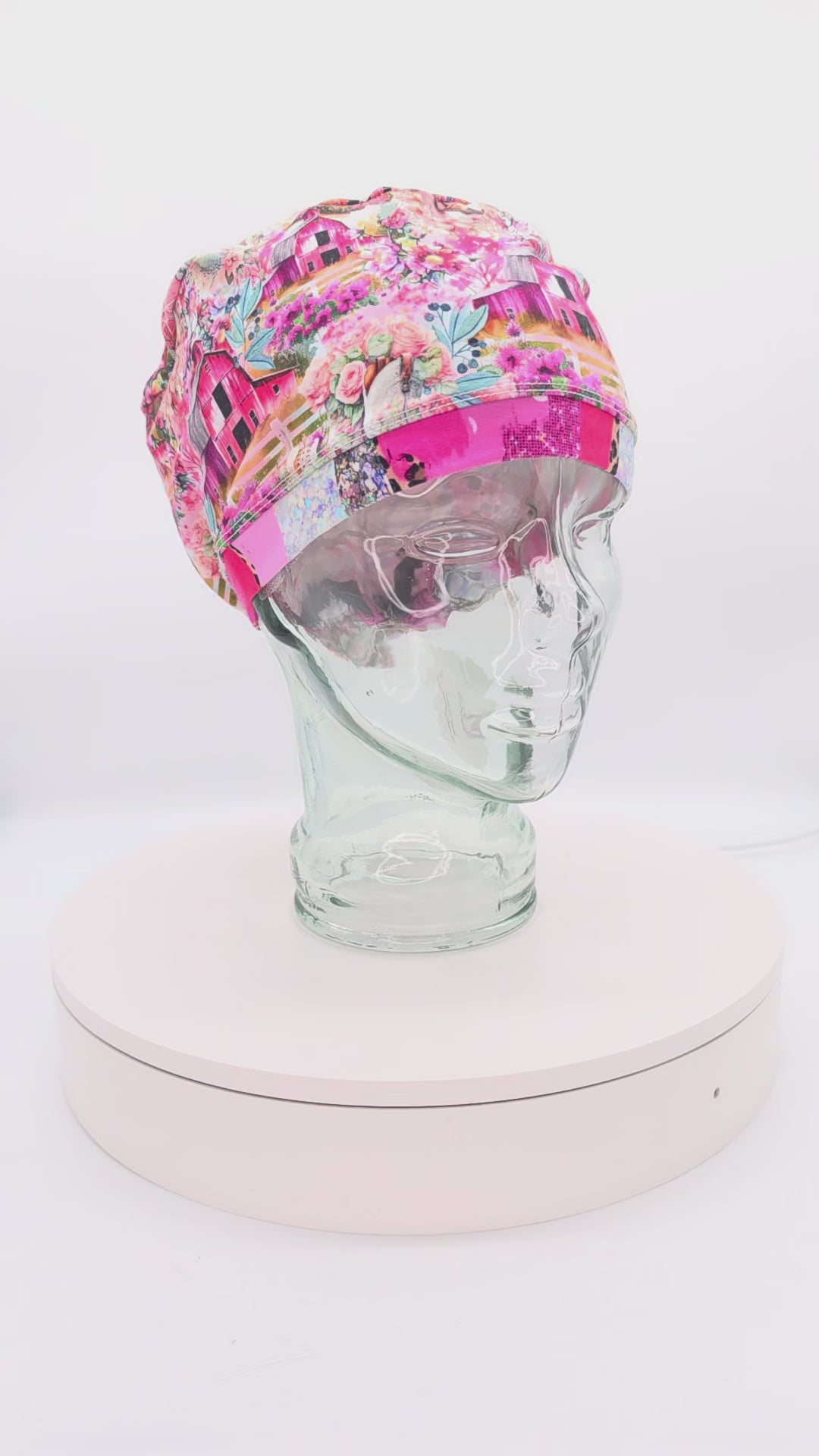 A 14-second rotating product video providing a 360-degree view of the Country Girl Full Coverage Scrub Cap on a clear mannequin head. The medical hat features a vibrant pink fabric patterned with a rustic Western floral and cowgirl motif, finished with a matching solid pink brim.