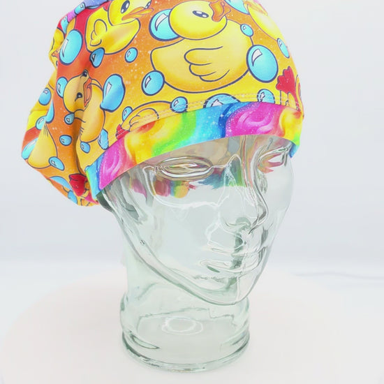 A 360-degree rotating view of a full coverage medical scrub cap on a mannequin, featuring a vibrant yellow rubber duck and blue bubble pattern.