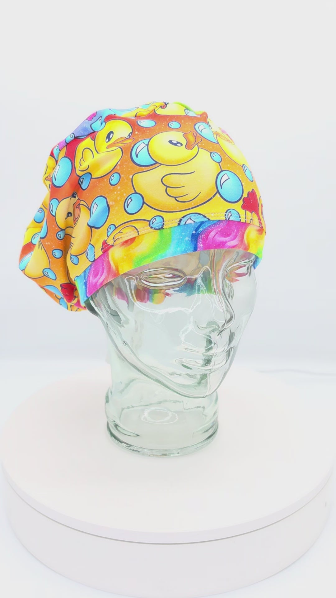 A 360-degree rotating view of a full coverage medical scrub cap on a mannequin, featuring a vibrant yellow rubber duck and blue bubble pattern.