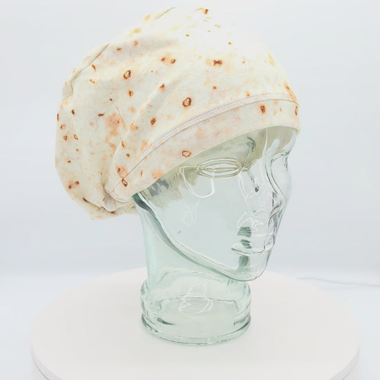 A 14-second rotating product video showing a 360-degree view of the Tortilla Full Coverage Scrub Cap displayed on a clear mannequin head. The medical hat features a hyper-realistic flour tortilla print with toasted brown spots and an adjustable black toggle closure in the back.