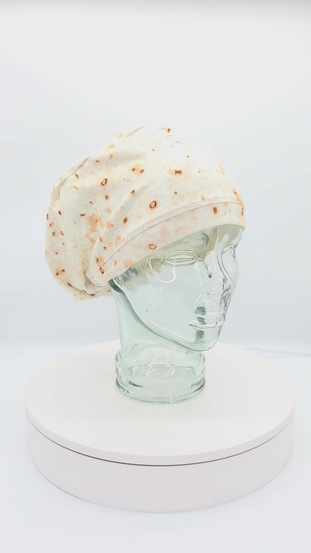 A 14-second rotating product video showing a 360-degree view of the Tortilla Full Coverage Scrub Cap displayed on a clear mannequin head. The medical hat features a hyper-realistic flour tortilla print with toasted brown spots and an adjustable black toggle closure in the back.