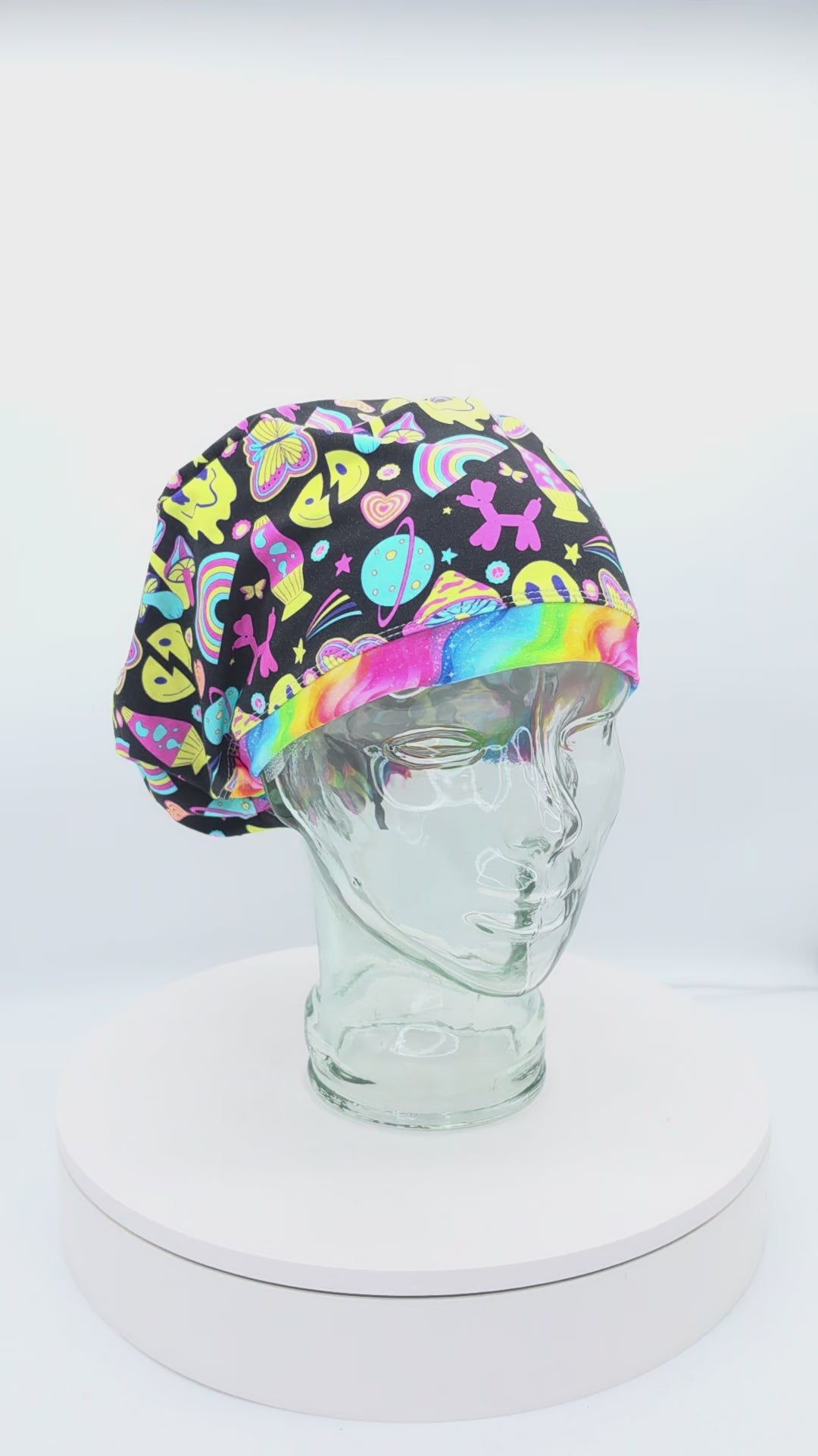 A 360-degree rotating video showcasing the Retro Days Scrub Cap on a clear mannequin head. This Euro-style medical hat features a vibrant, nostalgic 70s throwback print on a black fabric background filled with colorful smiley faces, rainbows, mushrooms, peace signs, and butterflies, accented by a rainbow swirl front brim.