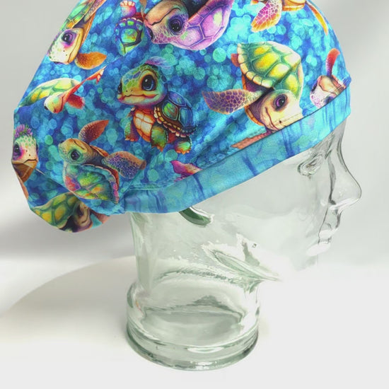 A 14-second product video featuring a handmade full-coverage scrub cap with a vibrant, colorful cartoon baby sea turtle pattern displayed on a clear glass mannequin head, slowly showcasing the secure fit and unique fabric design from the side.