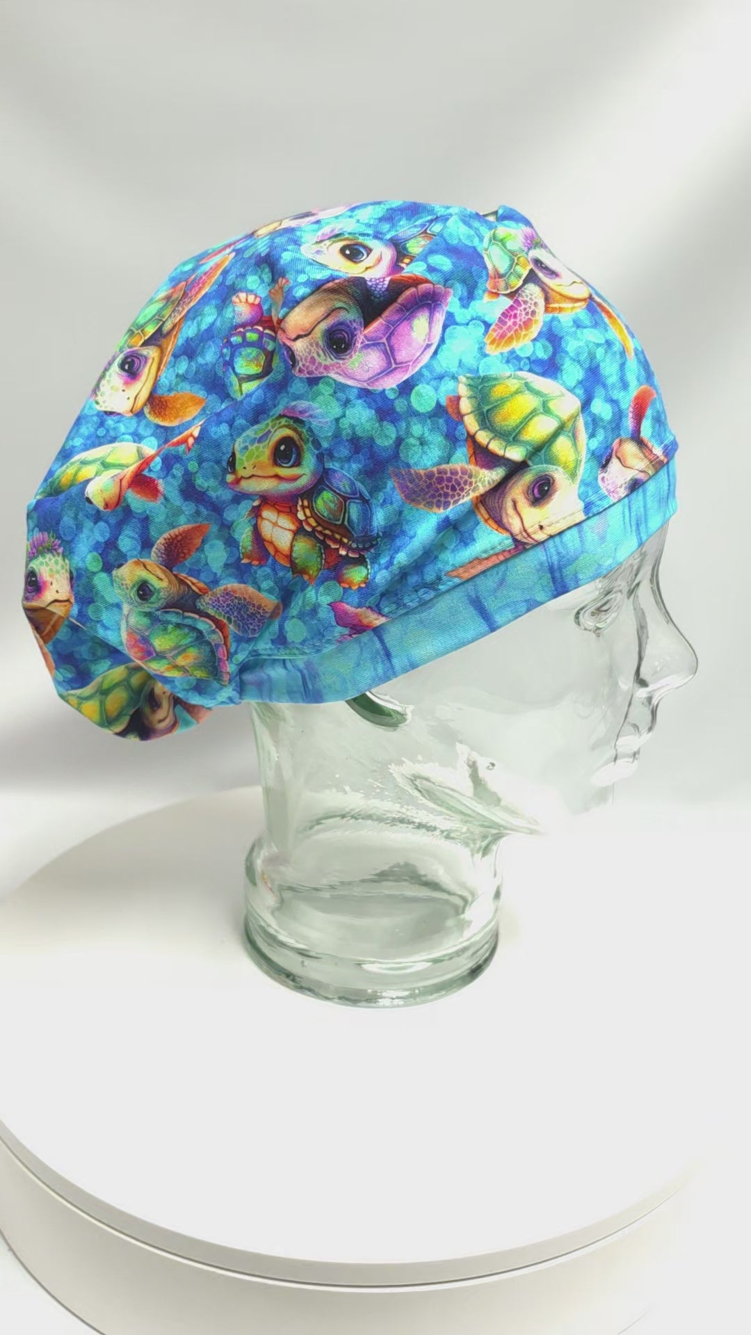 A 14-second product video featuring a handmade full-coverage scrub cap with a vibrant, colorful cartoon baby sea turtle pattern displayed on a clear glass mannequin head, slowly showcasing the secure fit and unique fabric design from the side.