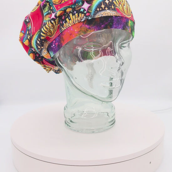 A 360-degree rotation video of a clear glass mannequin head on a white spinning display turntable, showcasing a colorful fast food scrub cap to demonstrate its full-coverage design and adjustable cord lock toggle fit from all angles.
