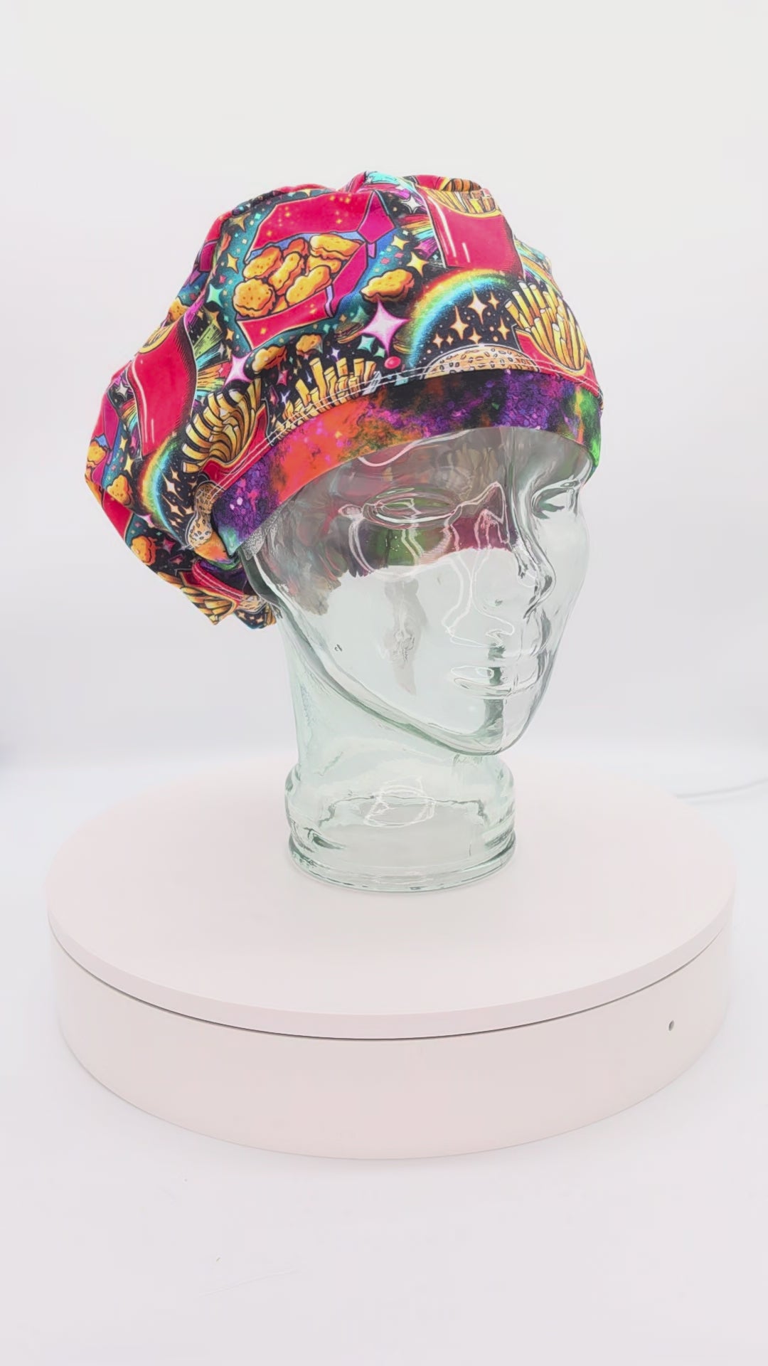 A 360-degree rotation video of a clear glass mannequin head on a white spinning display turntable, showcasing a colorful fast food scrub cap to demonstrate its full-coverage design and adjustable cord lock toggle fit from all angles.