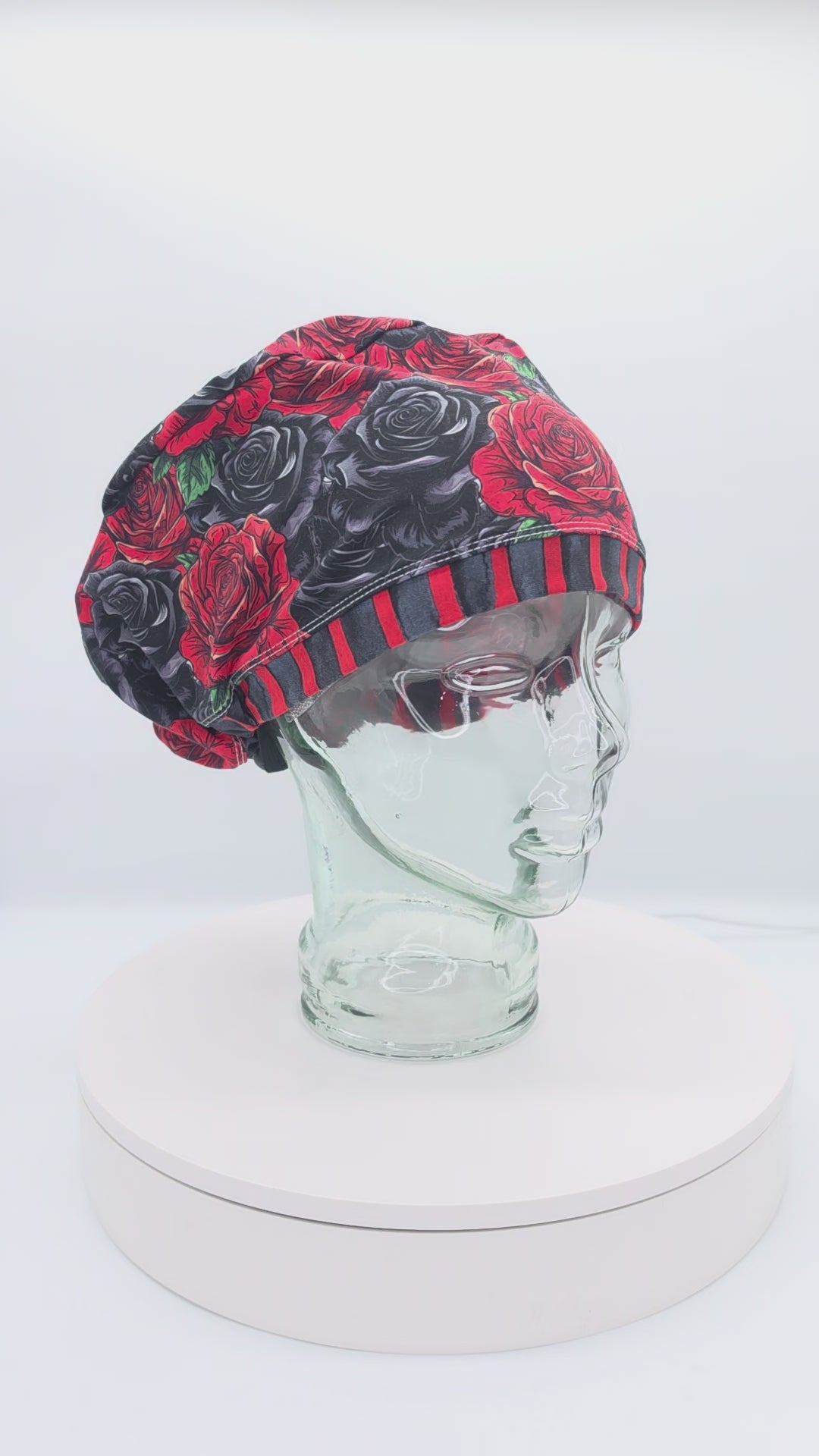 A 14-second rotating product video showing a 360-degree view of the Red and Black Roses Full Coverage Scrub Cap on a clear mannequin head. The medical hat features a dark gothic floral pattern of realistic red, charcoal, and black roses, finished with a bold red and black vertically striped brim.