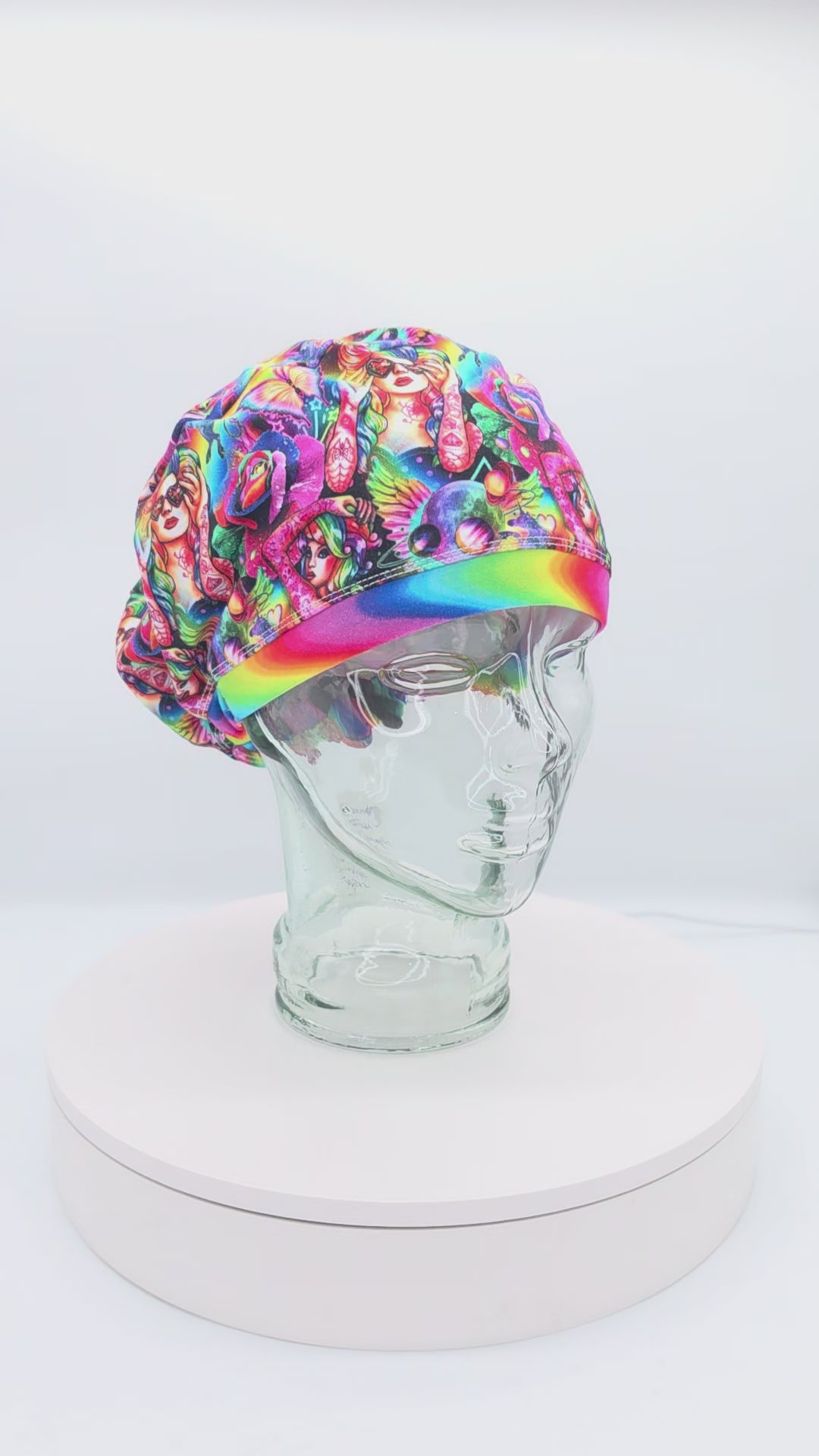 A 360-degree rotating view of the Rainbow Woman Euro-style scrub cap on a glass mannequin head, showcasing its vibrant, multicolored abstract face pattern against a black background and the adjustable back toggle closure.