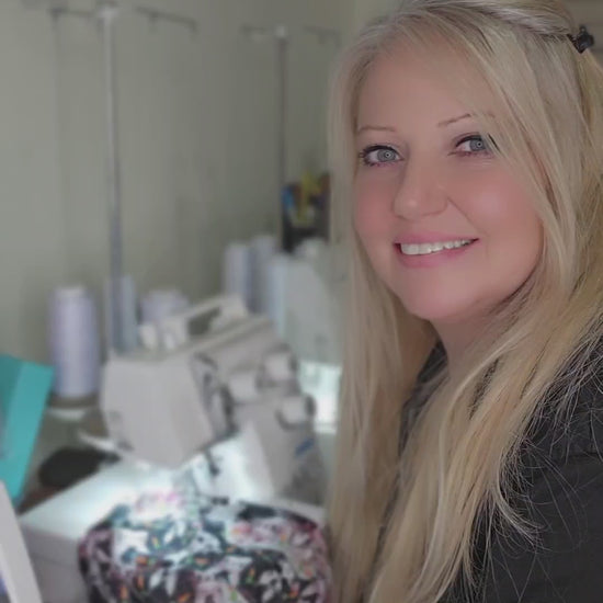 A behind-the-scenes look at the creation of a full-coverage medical scrub cap. A smiling blonde woman stands in her sewing studio, showing the premium fabric and handcrafted assembly process of the printed headwear, with sewing machine equipment visible in the background.