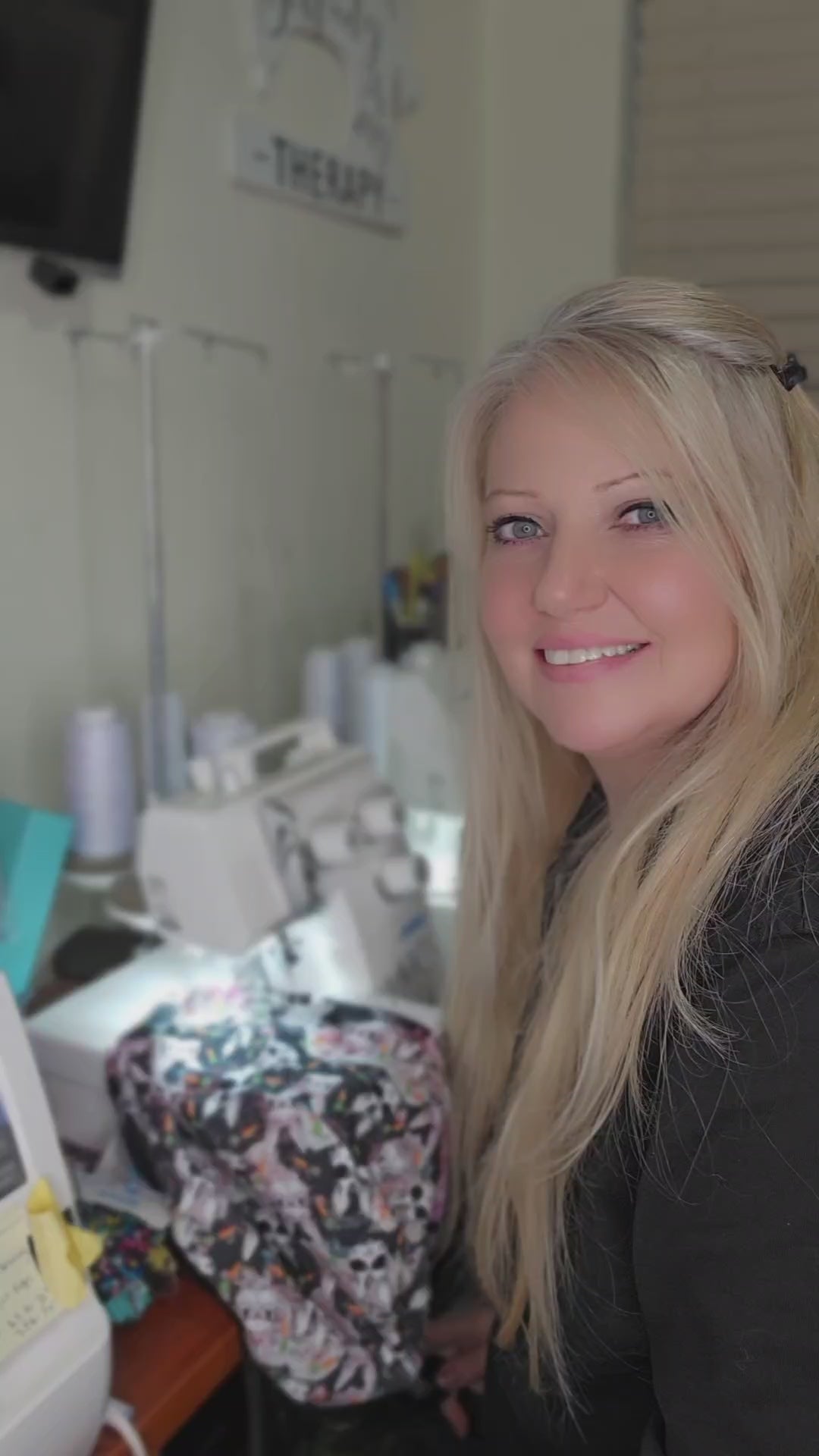A behind-the-scenes look at the creation of a full-coverage medical scrub cap. A smiling blonde woman stands in her sewing studio, showing the premium fabric and handcrafted assembly process of the printed headwear, with sewing machine equipment visible in the background.