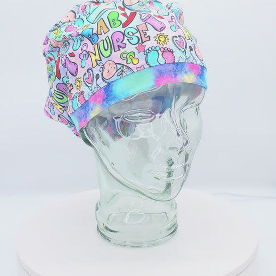 360-degree rotating view of a glass mannequin wearing a colorful "Baby Nurse" themed full-coverage Euro scrub cap. The print features vibrant bubble letters spelling "Baby Nurse" along with footprints, safety pins, and hearts, demonstrating the adjustable fit and secure back toggle.