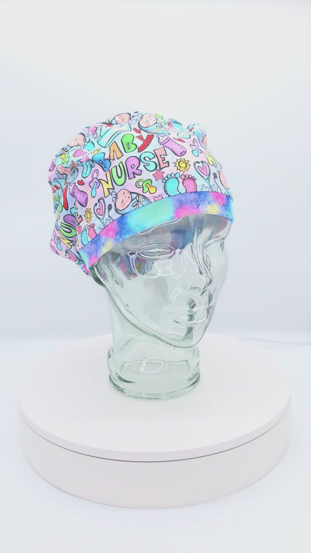 360-degree rotating view of a glass mannequin wearing a colorful "Baby Nurse" themed full-coverage Euro scrub cap. The print features vibrant bubble letters spelling "Baby Nurse" along with footprints, safety pins, and hearts, demonstrating the adjustable fit and secure back toggle.