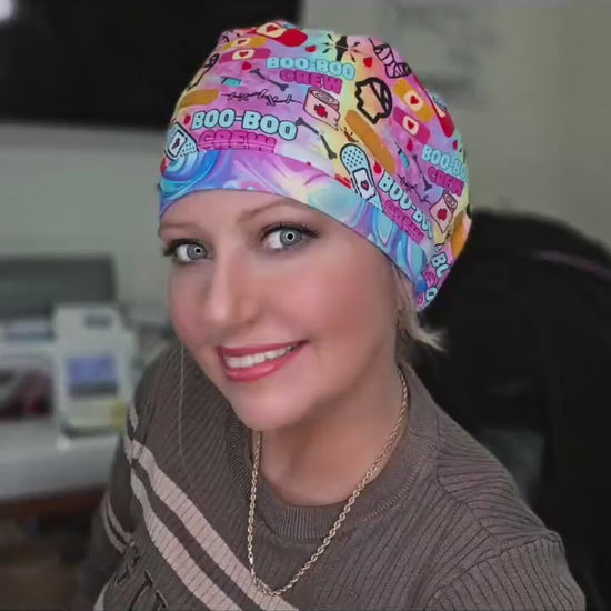 A 28-second video showcasing a smiling model wearing the colorful Boo Boo Crew full-coverage scrub cap. The cap features a vibrant, playful medical-themed print with cartoon bandages and "Boo-Boo Crew" text on a pink background.