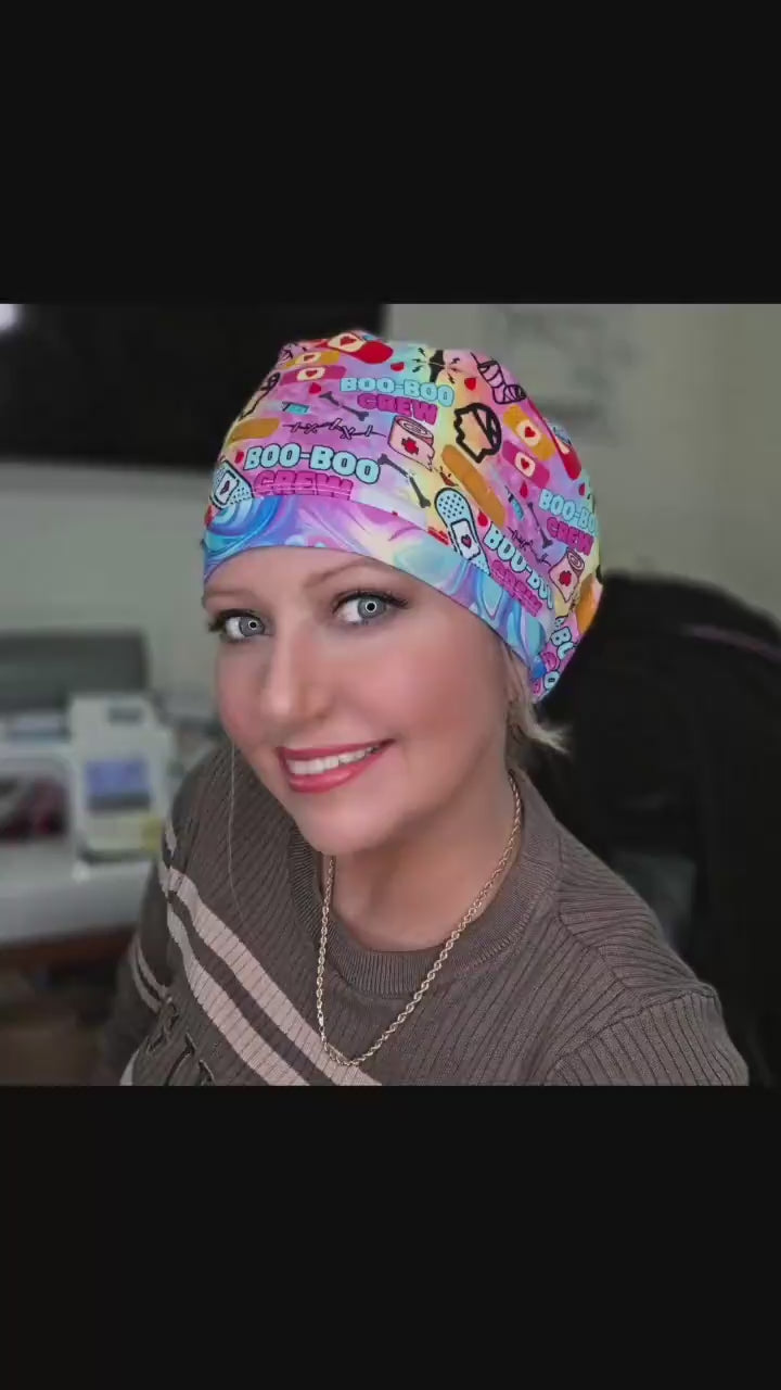 A 28-second video showcasing a smiling model wearing the colorful Boo Boo Crew full-coverage scrub cap. The cap features a vibrant, playful medical-themed print with cartoon bandages and "Boo-Boo Crew" text on a pink background.