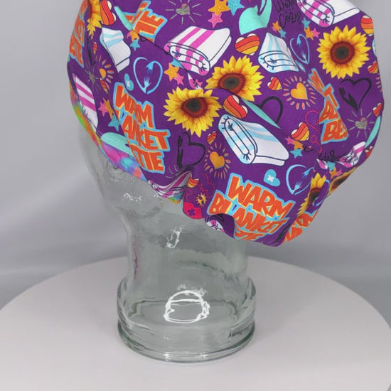 A 360-degree rotating video showcasing the Warm Blanket Bestie Euro-style scrub cap on a clear glass display head. The cap features a vibrant purple background covered in a detailed, colorful pattern with stylized sunflowers, red hearts, white and pink striped folded blankets, and bold text graphics reading "WARM BLANKET BESTIE" in orange and teal lettering. The video rotates to display the full coverage, contoured fit from all angles, highlighting the professional stitching and clean seams.