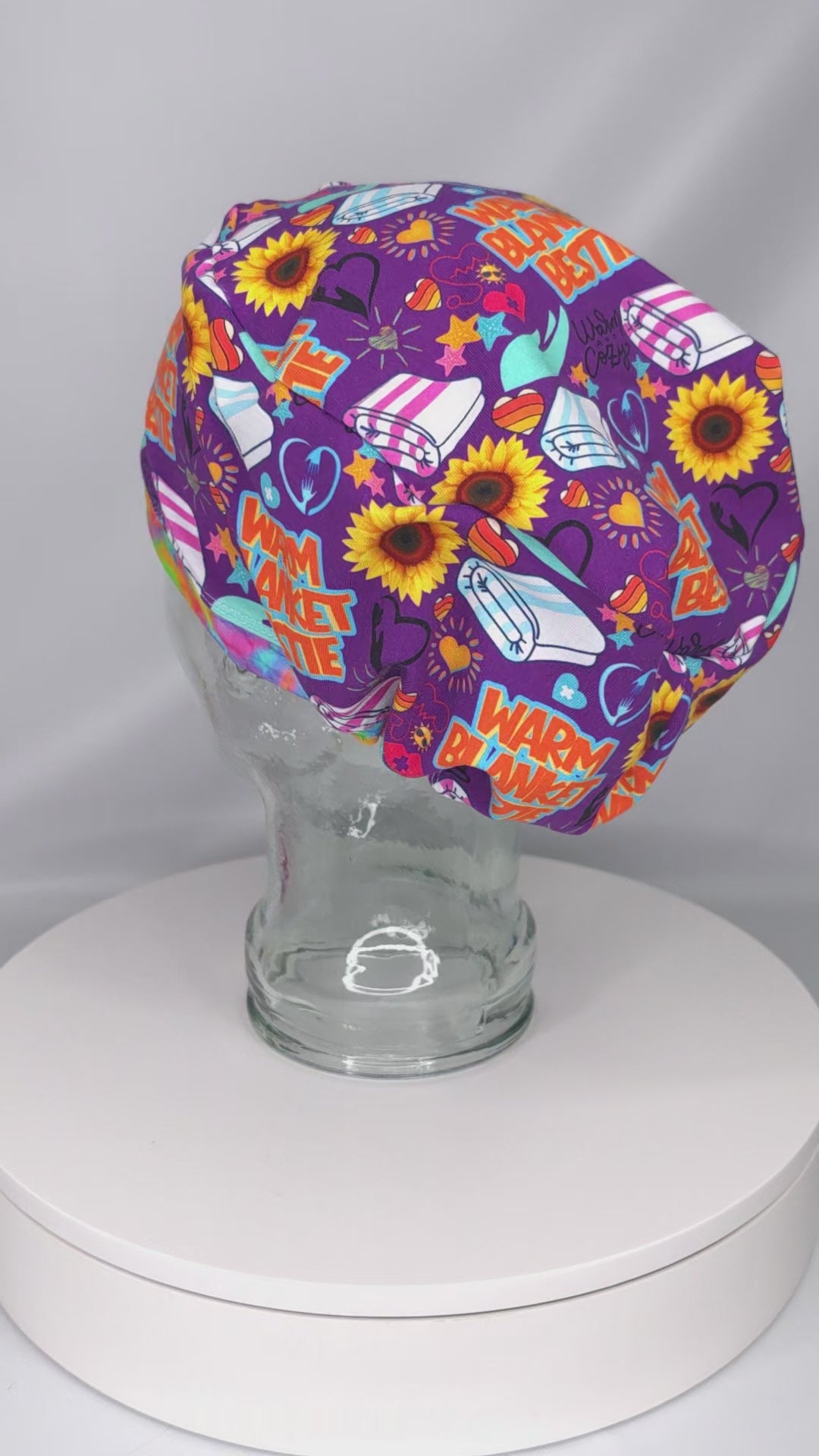 A 360-degree rotating video showcasing the Warm Blanket Bestie Euro-style scrub cap on a clear glass display head. The cap features a vibrant purple background covered in a detailed, colorful pattern with stylized sunflowers, red hearts, white and pink striped folded blankets, and bold text graphics reading "WARM BLANKET BESTIE" in orange and teal lettering. The video rotates to display the full coverage, contoured fit from all angles, highlighting the professional stitching and clean seams.