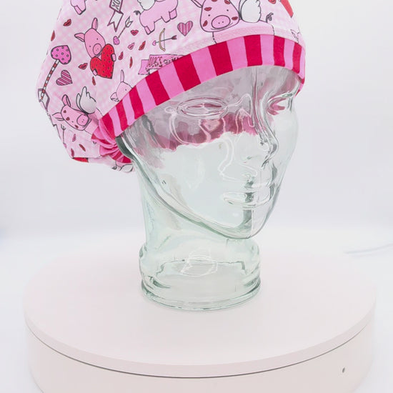 A 14-second rotating product video providing a 360-degree view of the Hogs and Kisses Full Coverage Scrub Cap on a clear mannequin head. The medical hat features a whimsical pink fabric patterned with cute illustrated cupids, cartoon pigs with wings, and floating hearts, finished with a contrasting red-and-pink striped brim.