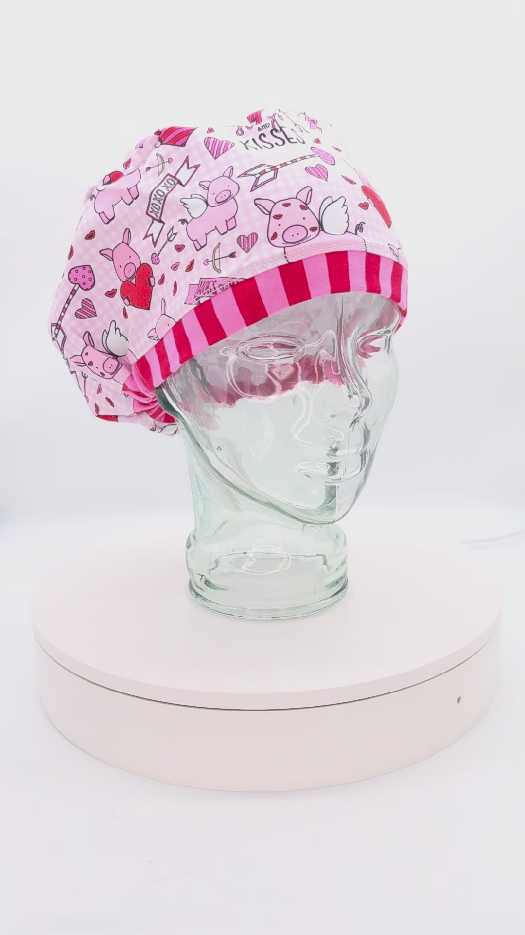 A 14-second rotating product video providing a 360-degree view of the Hogs and Kisses Full Coverage Scrub Cap on a clear mannequin head. The medical hat features a whimsical pink fabric patterned with cute illustrated cupids, cartoon pigs with wings, and floating hearts, finished with a contrasting red-and-pink striped brim.
