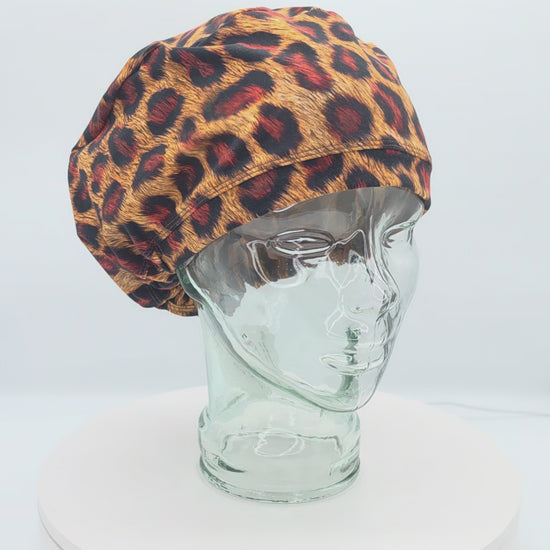 A 14-second rotating product video showing a 360-degree view of a clear mannequin head wearing the Leopard Cheetah Full Coverage Scrub Cap. The medical hat features a bold orange, red, and black leopard animal print pattern with a matching wide brim.