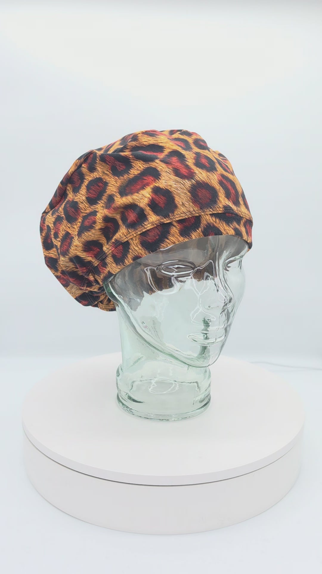 A 14-second rotating product video showing a 360-degree view of a clear mannequin head wearing the Leopard Cheetah Full Coverage Scrub Cap. The medical hat features a bold orange, red, and black leopard animal print pattern with a matching wide brim.